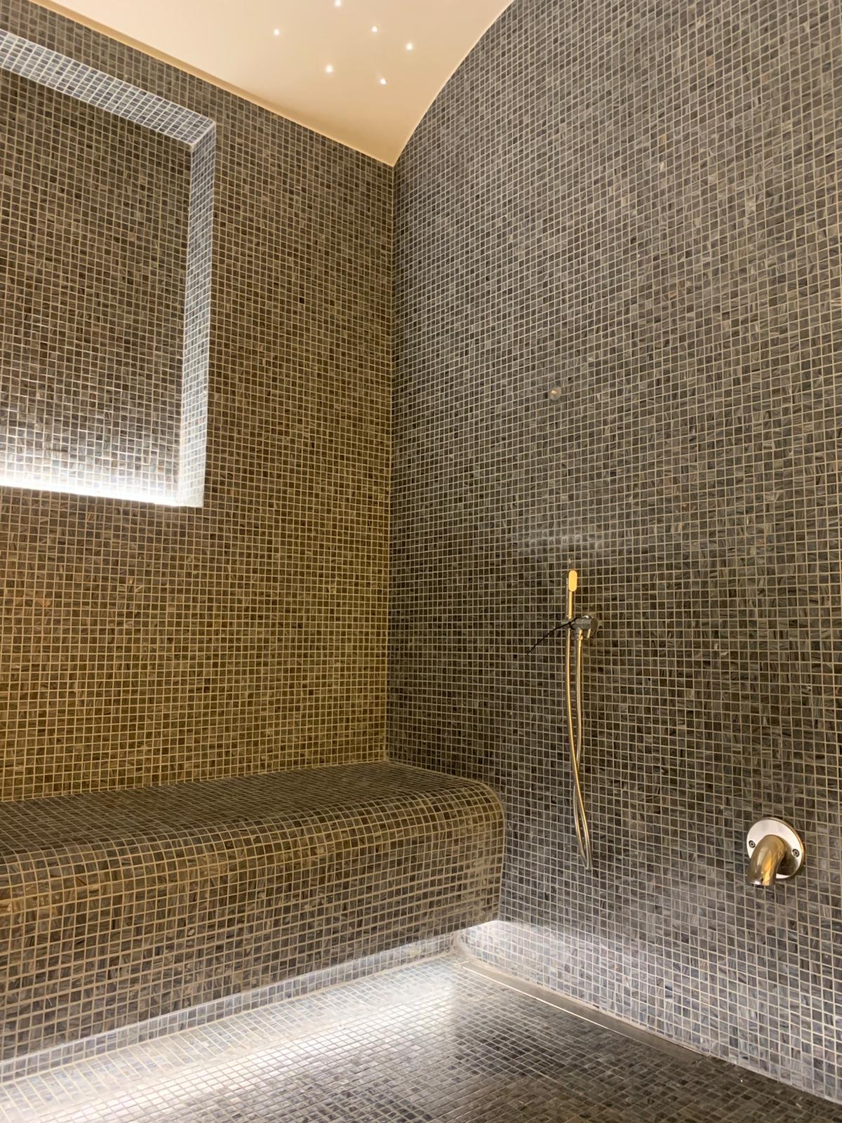 Steam room in a private villa in Dubai: dark grey glass mosaic on the walls and a curved bench, a ceiling that curves down into the wall, a lit mosaic niche, a hand shower and a steam outlet