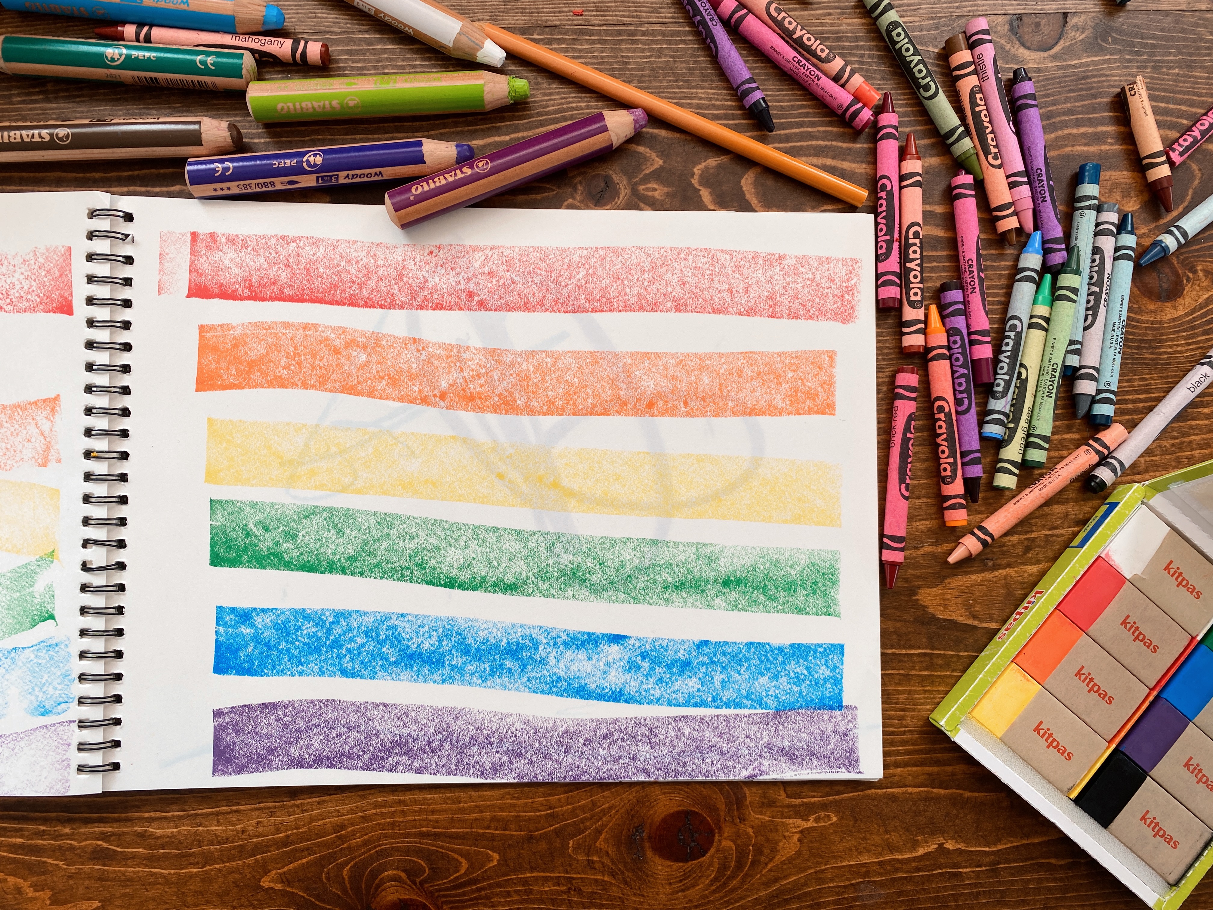 Wide rainbow stripes made with rectangular crayons make a Pride flag on a sketchbook page.