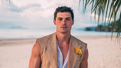 A man in a beige sleeveless blazer and light shirt stands on a beach