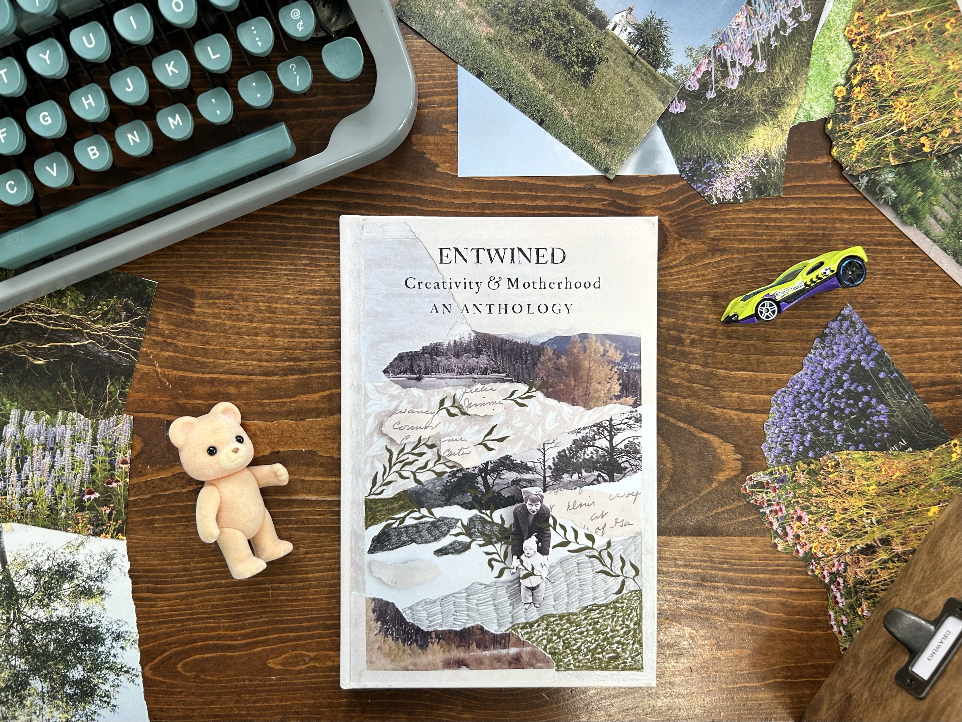 Entwined Creativity and Motherhood hardcover sitting on a desk with a teal typewriter, collage papers, toy bear and hot wheels car.