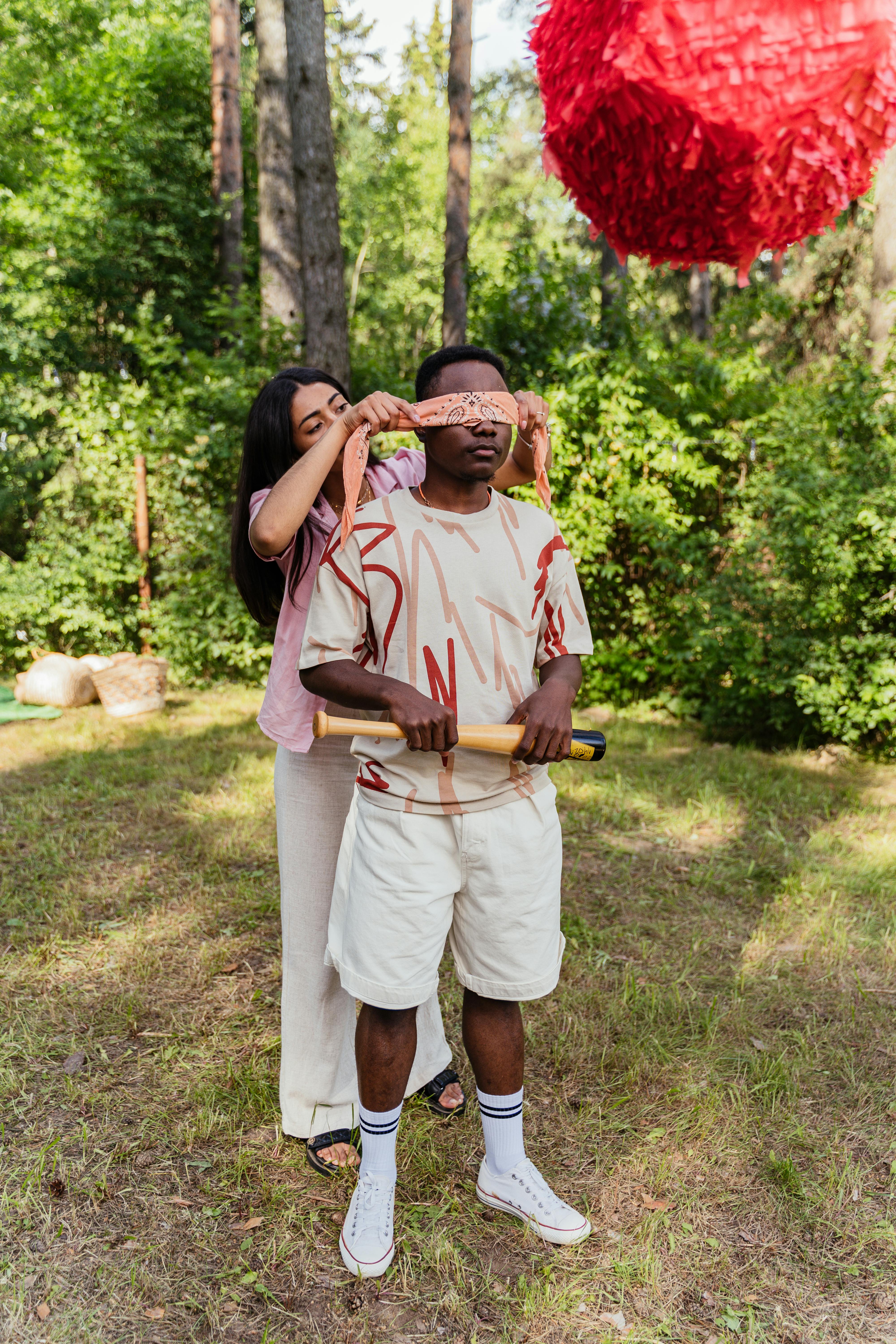 A man being blindfolded while holding a bat in a garden. There is a pinata in the foreground.