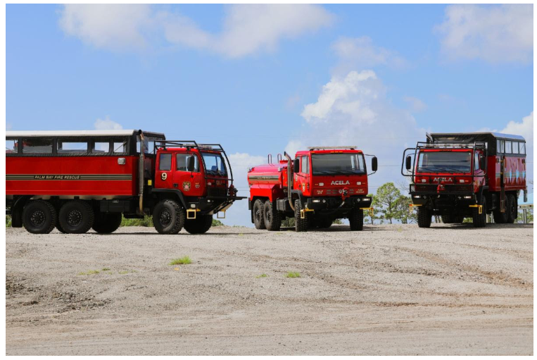Three red Palm Bay Fire Rescue Acela Monterra trucks parked on a gravel lot under a blue sky.