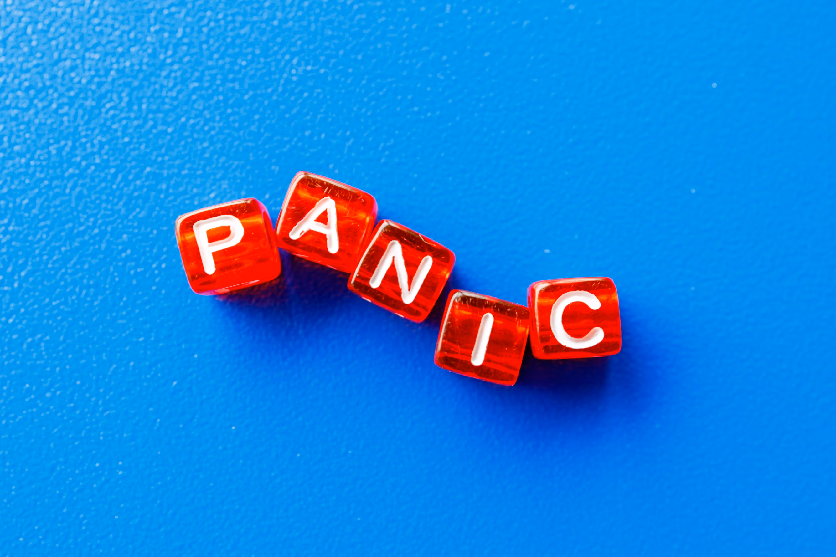 A picture from above of six red dice-like cubes with white letters on them, arranged to spell out the word "panic" in a lopsided but readable way. They are sitting on top of a blue textured background of some sort.