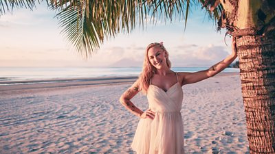 A woman in a white dress leans against a palm tree on a beach