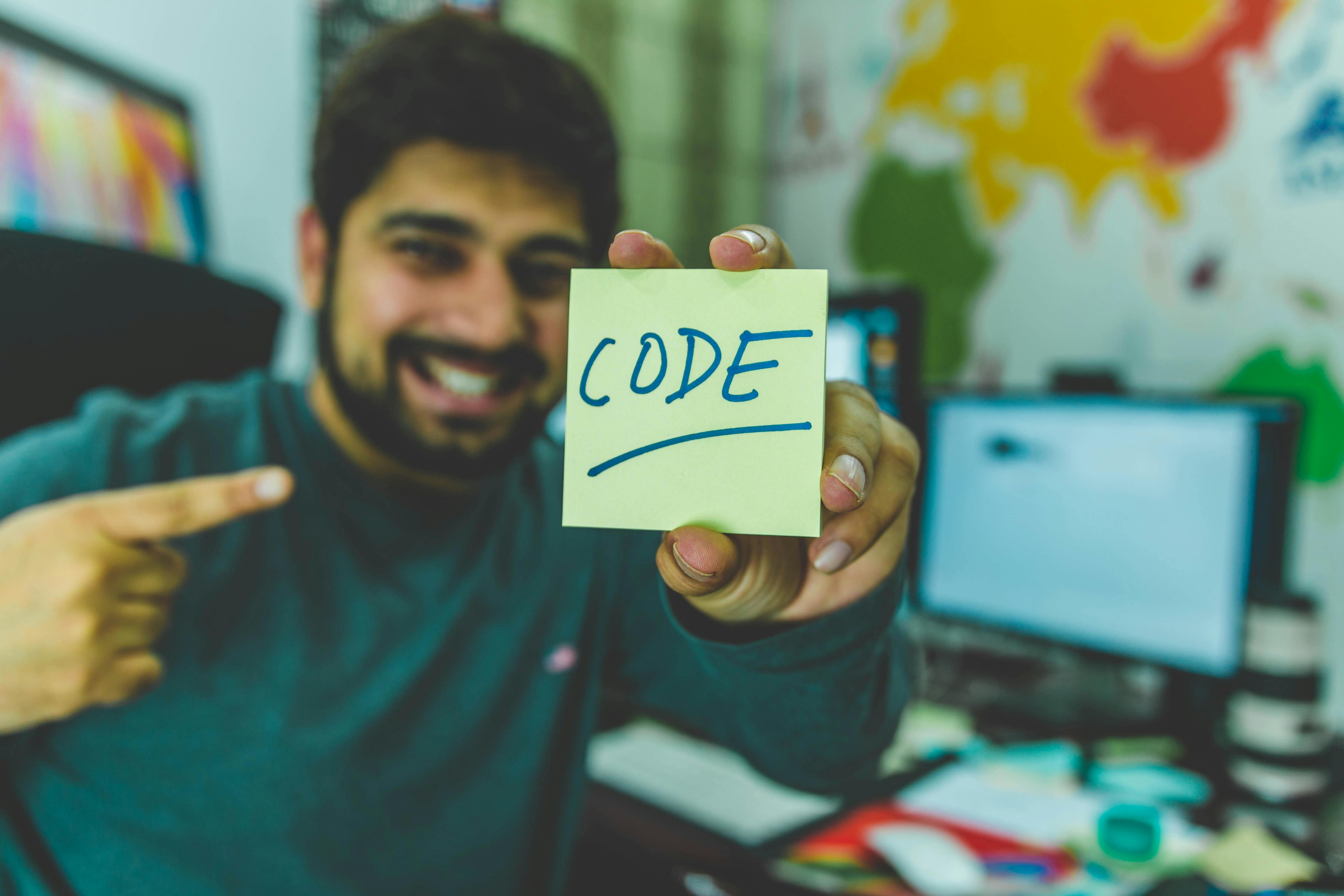 A smiling man holding and pointing at a post it note that says "code".