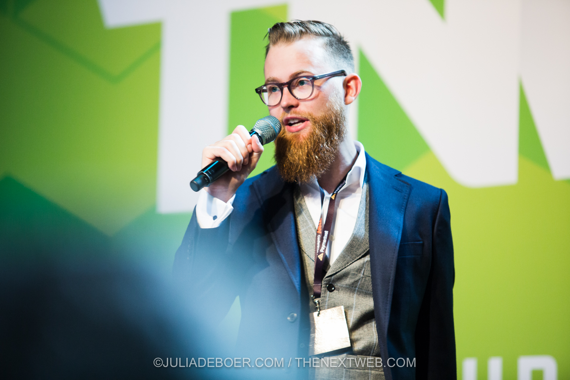 A bearded man with glasses is speaking into a handheld microphone, appearing to address an audience. He is dressed formally in a dark blazer over a light-colored vest and white shirt, with a lanyard around his neck holding an identification badge. The background features large green and white geometric shapes, suggesting a professional or conference setting.