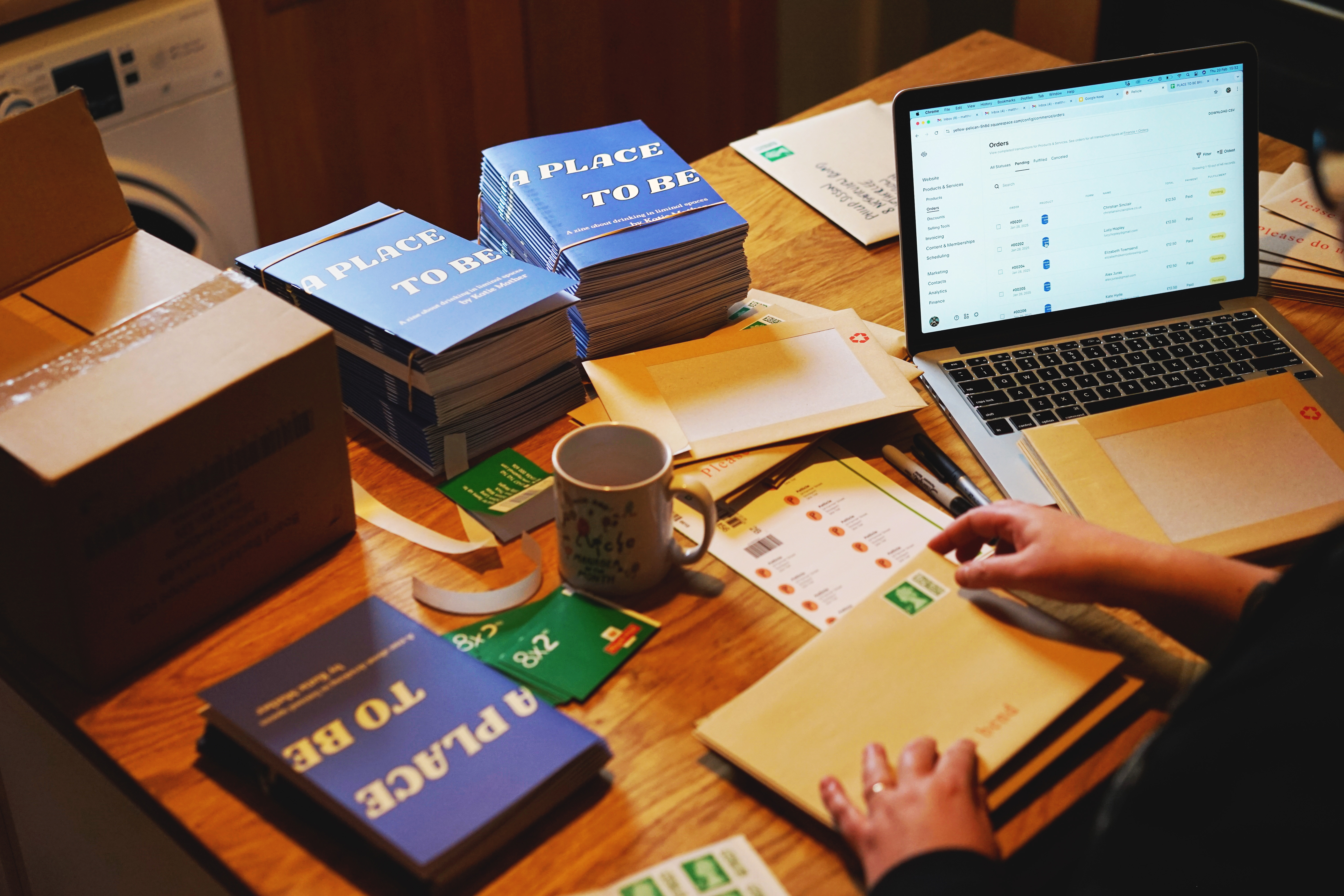 Packing A Place To Be zines at a kitchen table. Stacks of blue zines, green books of stamps, boxes of brown envelopes and an open laptop.