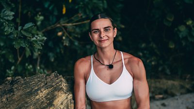 A woman in a white sports top smiles as she sits beside on a log on a tropical island