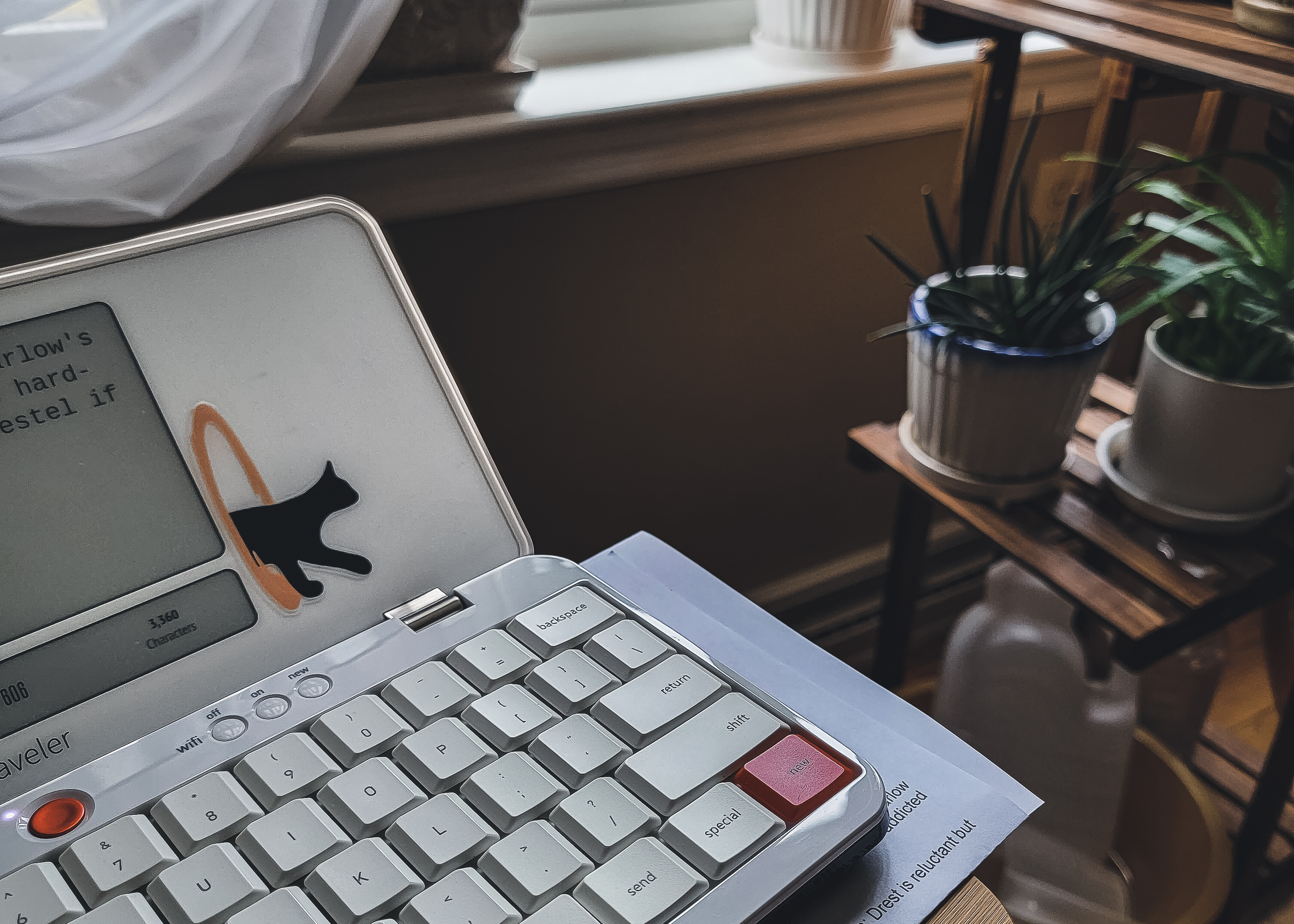 My Freewrite Traveler device with its white keyboard and e-ink screen sitting on the arm of my couch. A plant stand and windowsill are in the background.