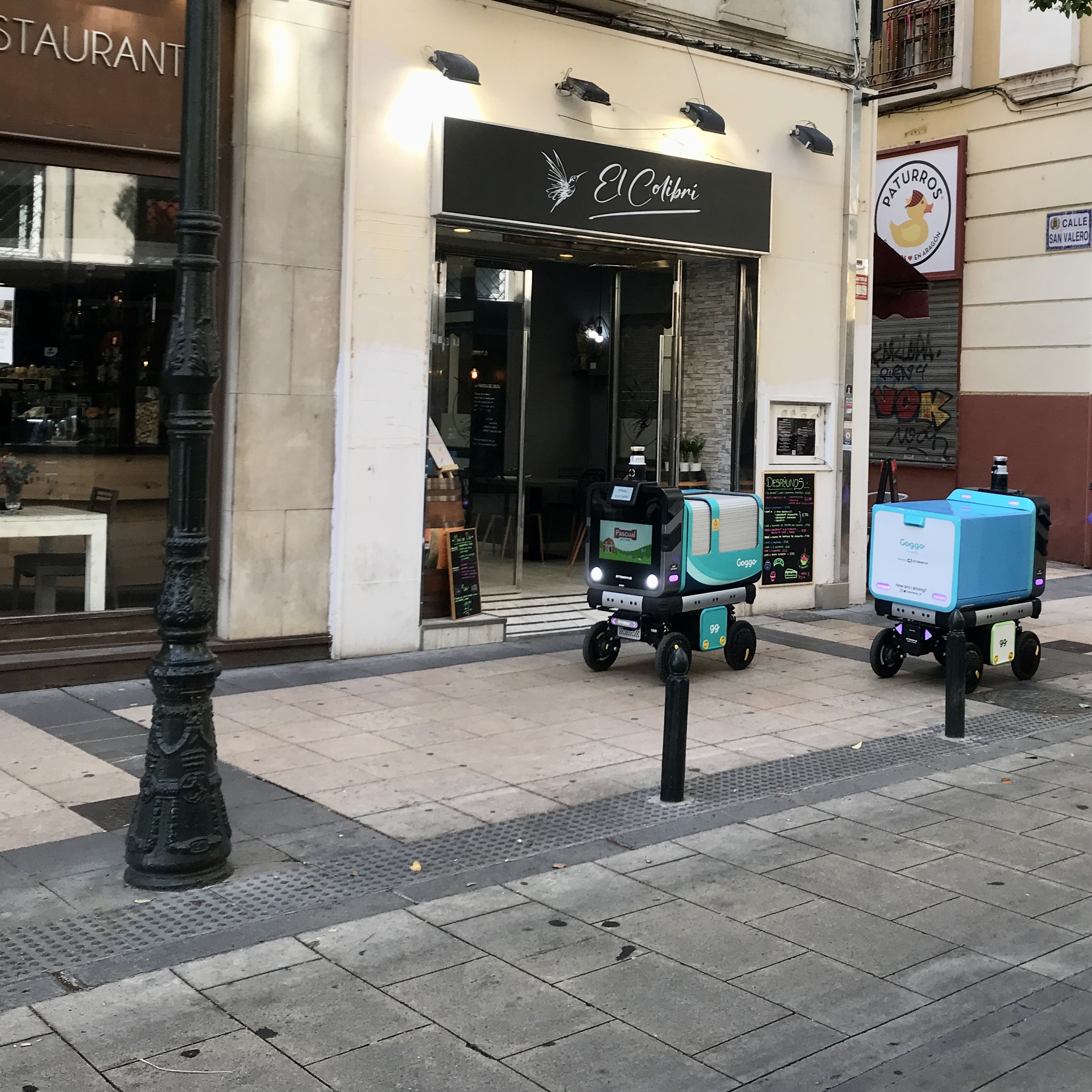 Dos robots de reparto en pruebas se cruzan en una acera de una calle semipeatonal impidiendo el paso a peatones