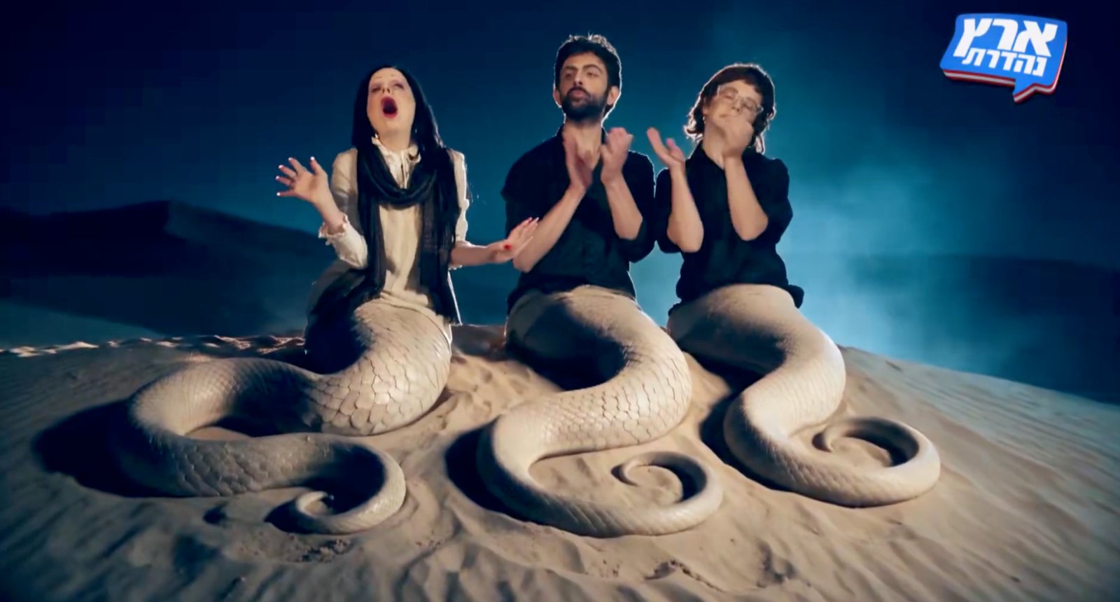 On Eretz Nehederet, three performers as half-human, half-snake sirens on a sand dune, one singing while two applaud.