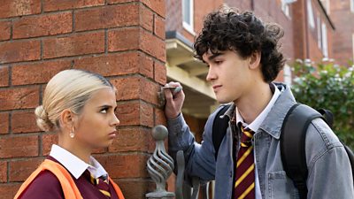 A girl and boy stand together by a wall looking tense