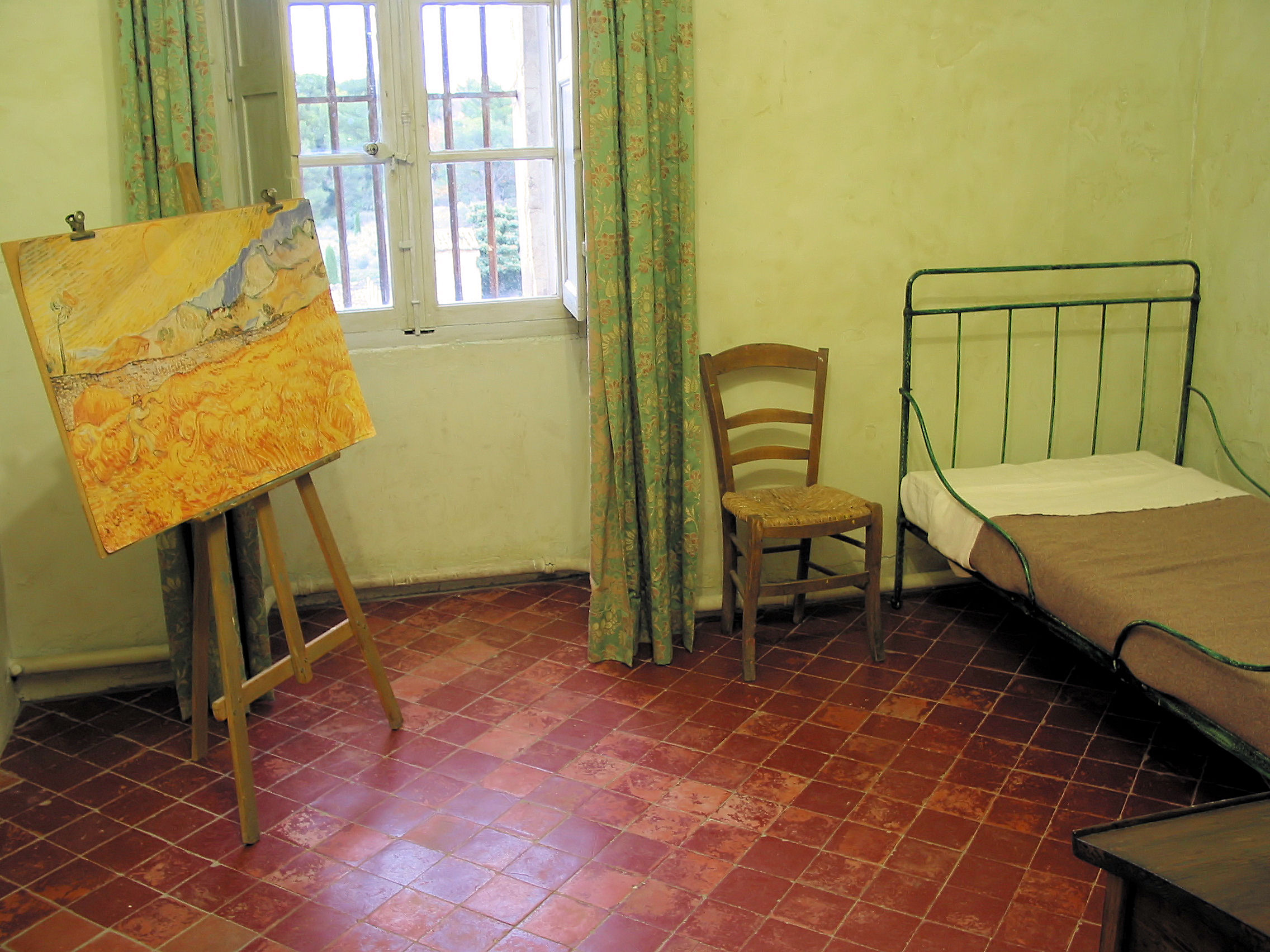 Simple room with a bed, chair, window, and easel holding a yellow painting